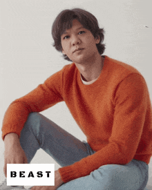 a man in an orange sweater sits on the floor with a beast logo behind him