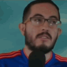 a man with glasses and a beard is wearing a blue shirt and talking into a microphone .