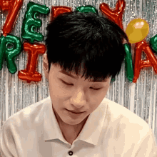 a young man in a white shirt is sitting in front of a wall with balloons and a curtain .