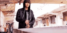 a young man wearing a leather jacket and a hoodie is standing on a sidewalk .