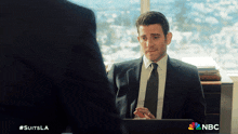 a man in a suit and tie is sitting in front of a laptop with nbc written on the bottom right