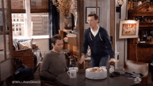 two men are sitting at a table in a living room with a cake on the table .