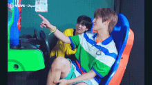 two young men are sitting in an arcade machine and one is pointing .