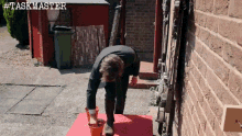 a man is pouring something into a bucket in front of a brick wall with # taskmaster written on the bottom