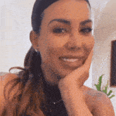 a woman wearing earrings and a necklace smiles with her hand on her chin