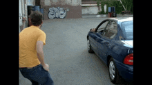 a man in a yellow shirt stands in front of a blue car with graffiti on the wall that says snc