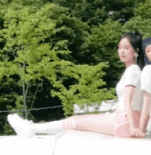 two women are sitting on a roof with trees in the background . one of the women is wearing knee high socks .