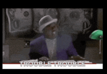 a man in a purple suit and white hat is sitting at a desk with the words trouble trouble on the bottom