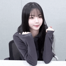 a girl with long black hair is sitting at a desk with her hands on her chin