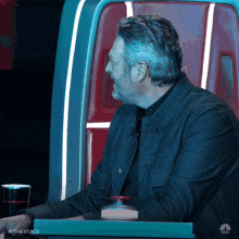 a man is sitting in a chair with a red button in front of him that says the voice