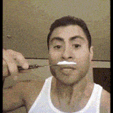 a man in a white tank top is shaving his beard with a razor