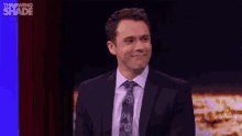 a man in a suit and tie is sitting on a stage and saying `` thank you '' .