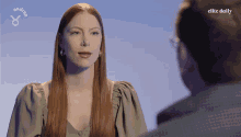a woman with long red hair talks to a man in front of a blue background that says elite daily