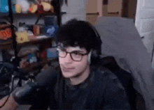 a young man wearing glasses and headphones is sitting in front of a microphone in a bedroom .