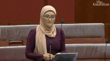 a woman wearing a hijab and glasses is giving a speech in front of a microphone .