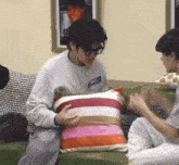 a man is sitting on a couch holding a striped pillow while another man looks on .