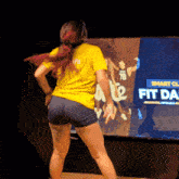 a woman in a yellow shirt is dancing in front of a screen that says " fit day "