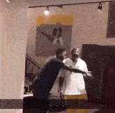 two men are standing in a room with a picture on the wall behind them