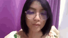 a woman wearing glasses is standing in front of a purple curtain and making a funny face .