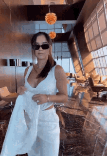 a woman wearing sunglasses and a white top is standing in a room with a lot of windows .