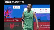 a man wearing a green petronas shirt stands on a badminton court