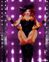 a drag queen is standing in front of a purple curtain with lights behind her