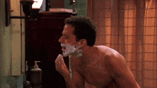 a shirtless man shaves his face in front of a mirror with a bottle of soap on the counter