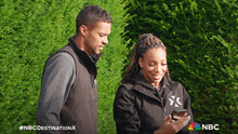 a man and a woman are looking at a cell phone with the nbc destination x written on the bottom