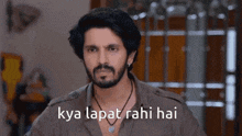 a man with a beard is standing in front of a door with the words `` kya lapit rahi hai '' written on the screen .