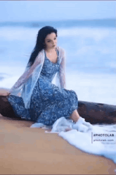 a woman in a blue dress is sitting on a log on a beach