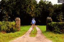 a man is walking down a dirt road with buzza written on the bottom right
