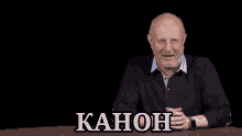 a bald man with glasses and a beard is sitting at a table with the word kahon written on it