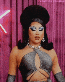 a drag queen is standing in front of a pink curtain wearing a silver top and gloves