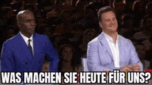 two men in suits and ties are sitting in front of a crowd with the words was machen sie heute for uns written above them .