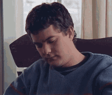 a young man in a blue sweater is sitting in a chair looking down .
