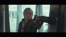 a man is holding a gun in front of a window and pointing it at the camera .