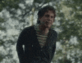 a man in a hoodie and striped shirt is standing in the woods with his hands in his pockets .