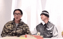 two men are sitting at a table and one has a beanie that says " hungry "