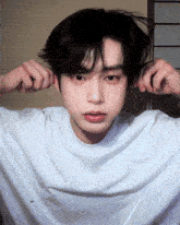 a young man wearing a white t-shirt has his hands on his ears looking at the camera