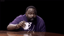 a man in a purple shirt is sitting at a table drinking a cup of coffee .