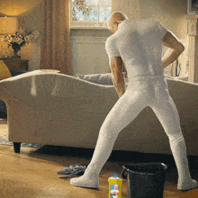 a man in white pants is standing in a living room next to a bucket and a bottle of mr. clean