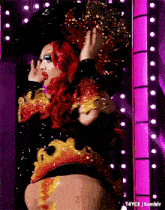 a drag queen with red hair is standing in front of a pink wall with lights behind her
