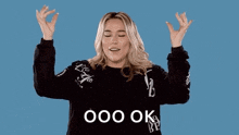 a woman in a black sweater is holding her hands up in the air and saying `` ooo ok '' .