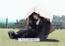 a woman in a black dress is sitting on the grass with a cardboard box on her head