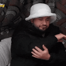 a man with a beard wearing a white hat and a black jacket is sitting on a couch .