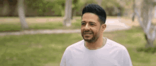 a man with a beard wearing a white t-shirt is smiling in a park .