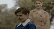 a young boy in a blue shirt is standing next to a woman in a trench coat .