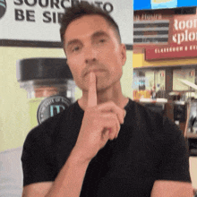 a man in a black shirt holds his finger to his lips in front of a sign that says source to be sir