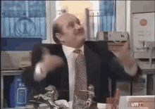 a bald man in a suit and tie is sitting at a desk with his arms outstretched .