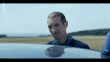 a man in a blue shirt is standing in front of a blue car with a reflection of him in the hood